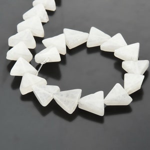 May include: A string of white, triangular gemstone beads. Each bead is a translucent, milky white colour. The beads are strung together, creating a necklace or bracelet component. The background is a dark, solid colour.