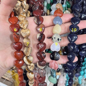 May include: A close-up of ten strands of heart-shaped gemstone beads. The beads are in various colors, including red, brown, white, blue, and black. The beads are arranged in a line, with the numbers 6, 7, 8, 9, and 10 written on the beads.