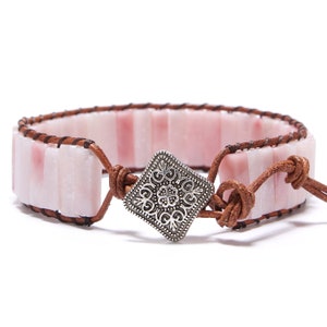 Könnte beinhalten: Ein Wickelarmband mit rechteckigen rosa Steinen und braunem Leder. Das Armband wird mit einem dekorativen quadratischen Verschluss in Silberfarbe befestigt. Das Leder wickelt sich um das Handgelenk und schafft ein stilvolles Accessoire.