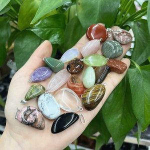 May include: A selection of polished gemstone pendants in diverse shapes and colours, including red, green, purple, and brown. Each stone has a silver loop for hanging. The pendants are held in a hand against a backdrop of green leaves.