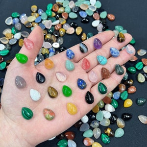 Puede incluir: Surtido de colgantes de piedra preciosa en forma de lágrima de colores, incluyendo verde, azul, rojo y marrón. Los colgantes están dispuestos en una mano, mostrando su tamaño y forma.