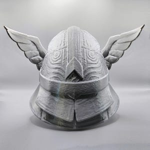 May include: A silver-colored helmet with wing-like structures on each side. The helmet has intricate details and appears to be made of metal. The design suggests a fantasy or historical theme.