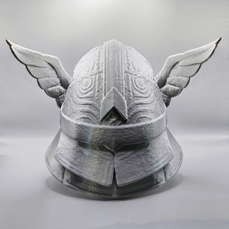 May include: A silver-colored helmet with wing-like structures on each side. The helmet has intricate details and appears to be made of metal. The design suggests a fantasy or historical theme.