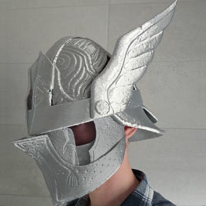 May include: A silver-colored helmet with a winged design on the side. The helmet has intricate details and a visor. The person wearing the helmet is wearing a plaid shirt.