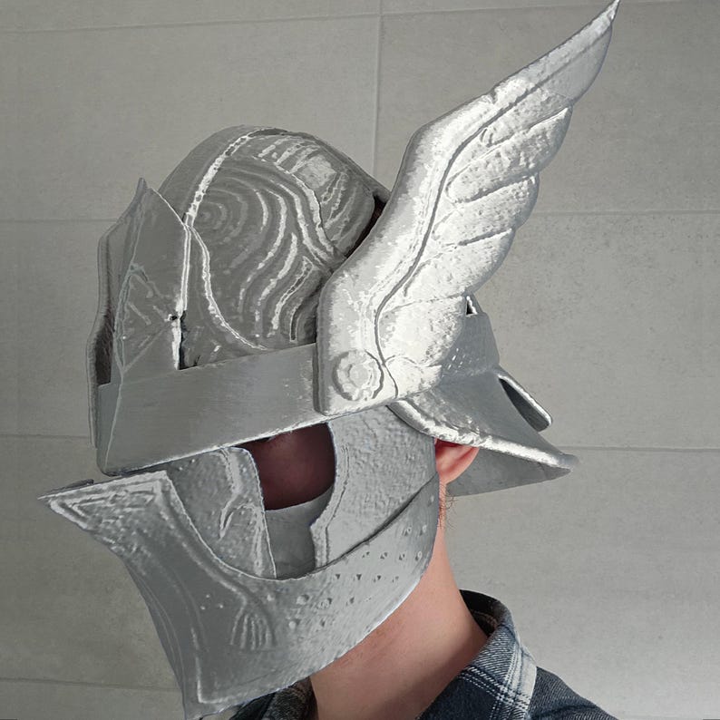 May include: A silver-colored helmet with a winged design on the side. The helmet has intricate details and a visor. The person wearing the helmet is wearing a plaid shirt.