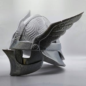 May include: A gray helmet with wing-like features on the sides. The helmet has a detailed design with a textured surface and a visor. The helmet appears to be made of a hard material.