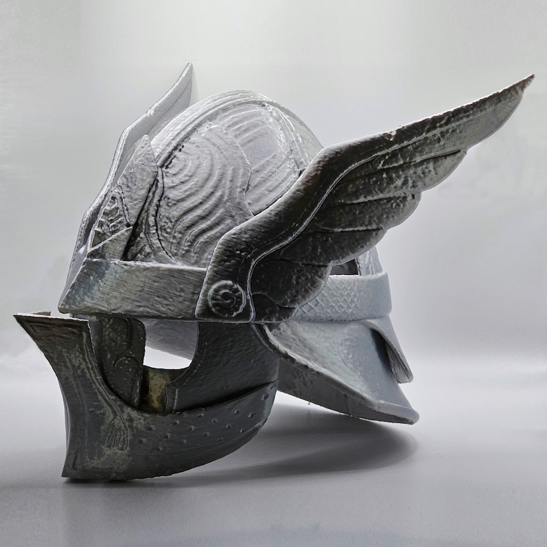May include: A gray helmet with wing-like features on the sides. The helmet has a detailed design with a textured surface and a visor. The helmet appears to be made of a hard material.