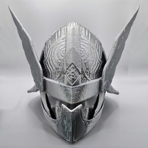 May include: A silver-colored helmet with wing-like protrusions on the sides. The helmet has intricate embossed patterns and a visor. The design suggests a historical or fantasy theme, possibly for cosplay or display.