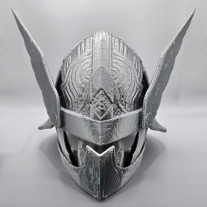 May include: A silver-colored helmet with wing-like protrusions on the sides. The helmet has intricate embossed patterns and a visor. The design suggests a historical or fantasy theme, possibly for cosplay or display.