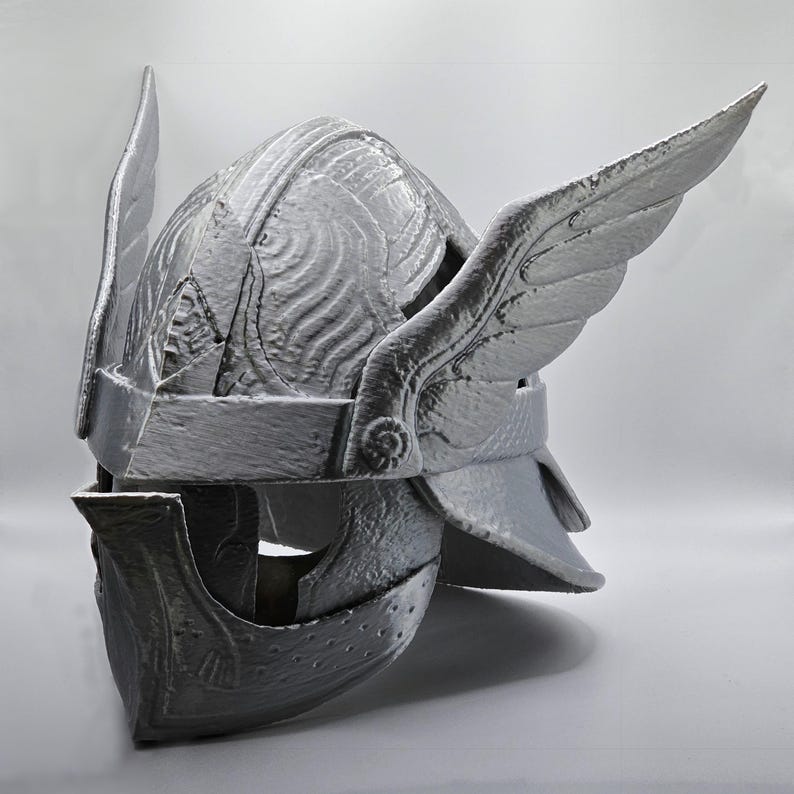 May include: A silver-colored helmet with wing-like features on the sides. The helmet has detailed carvings and a visor. The design suggests a historical or fantasy theme, possibly for cosplay or display.
