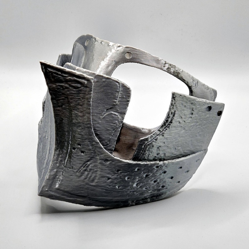 May include: A metallic gray, medieval-style mask with a curved top handle. The mask has a textured surface with small, evenly spaced holes and riveted details. The design suggests a historical or fantasy theme.