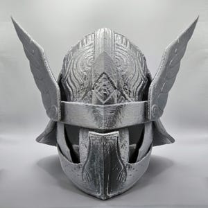 May include: A silver-colored helmet with wing-like protrusions on each side. The helmet features intricate embossed patterns and a visor. The design suggests a historical or fantasy theme, possibly for cosplay or display.