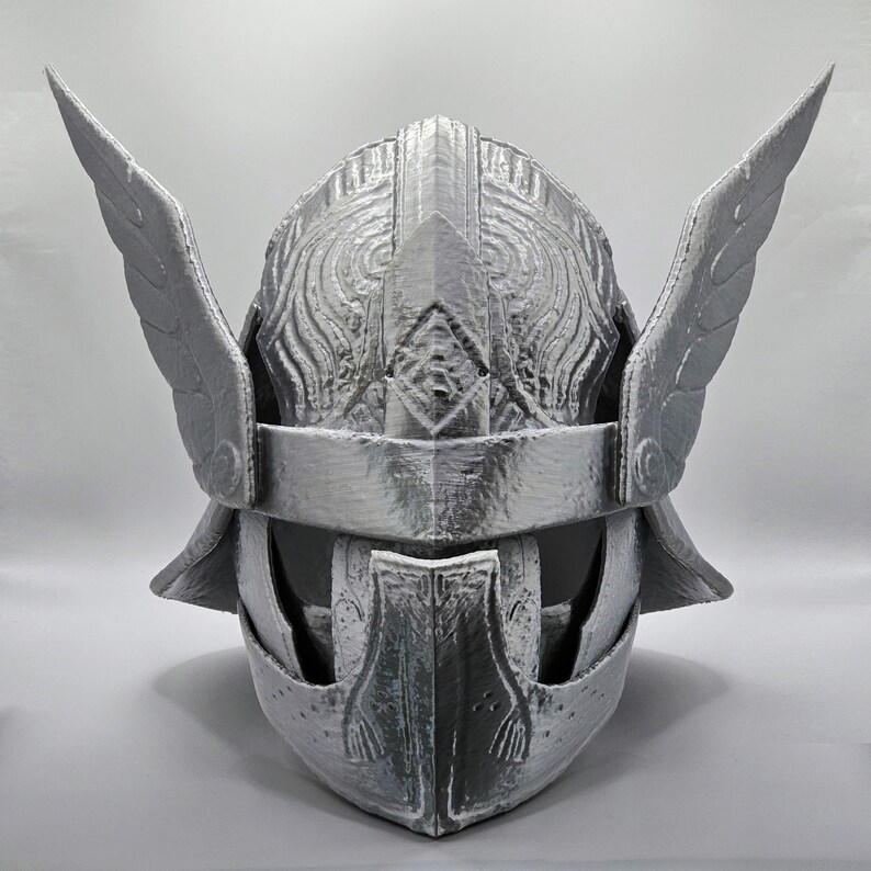 May include: A silver-colored helmet with wing-like protrusions on each side. The helmet features intricate embossed patterns and a visor. The design suggests a historical or fantasy theme, possibly for cosplay or display.