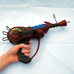 May include: A red and black ray gun with blue accents and a gauge labelled "Shot-O-Matic". The prop gun has a handle and a curved trigger guard. The barrel has a small red tip and a black antenna.