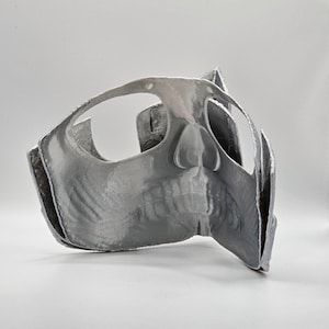May include: A gray, 3D-printed mask with a skull-like design. The mask features eye openings, a nose, and a jawline. The mask is made of a smooth, reflective material and is likely for costume or decorative purposes.