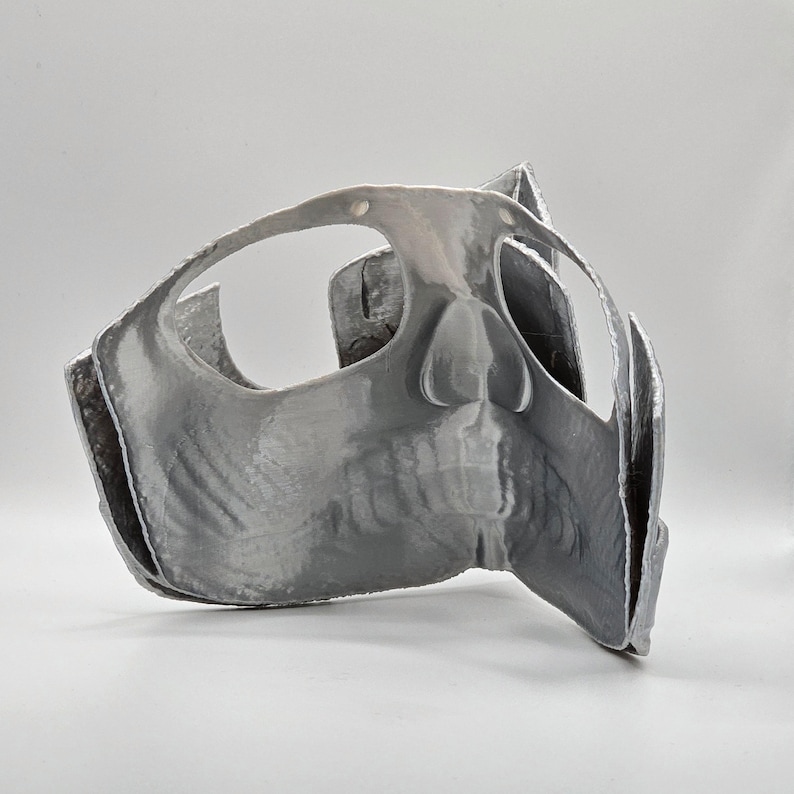 May include: A gray, 3D-printed mask with a skull-like design. The mask features eye openings, a nose, and a jawline. The mask is made of a smooth, reflective material and is likely for costume or decorative purposes.