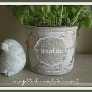 Puede incluir: Una maceta de cerámica blanca con un diseño decorativo y una etiqueta que dice "Jardin du Basilic Septembre". La maceta está llena de plantas de albahaca verde y está sentada sobre una superficie marrón claro. Una pequeña figurita de pájaro gris también está en la superficie.
