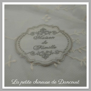 May include: A white fabric label with silver embroidery that reads "Maison de Famille". The label is on a white lace background.