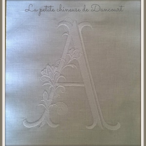 Può includere: Ricamo bianco della lettera "A" con dettagli floreali su tessuto chiaro. Il testo "La petite chineuse de Dancourt" è sopra la lettera. Il ricamo è dettagliato ed elegante, in stile monogramma classico.