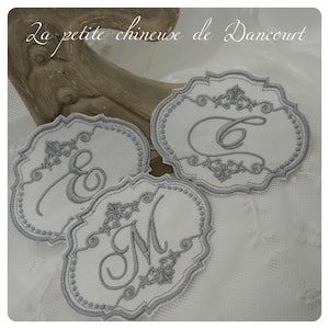 Peut inclure: Trois dessous-de-verre en tissu blanc avec des lettres brodées grises C, E et M. Les dessous-de-verre ont un bord festonné et un motif floral décoratif.