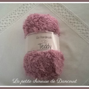 Puede incluir: Un ovillo de lana rosa con la etiqueta "Go Handmade Teddy" sobre una superficie blanca con ribete de encaje blanco.
