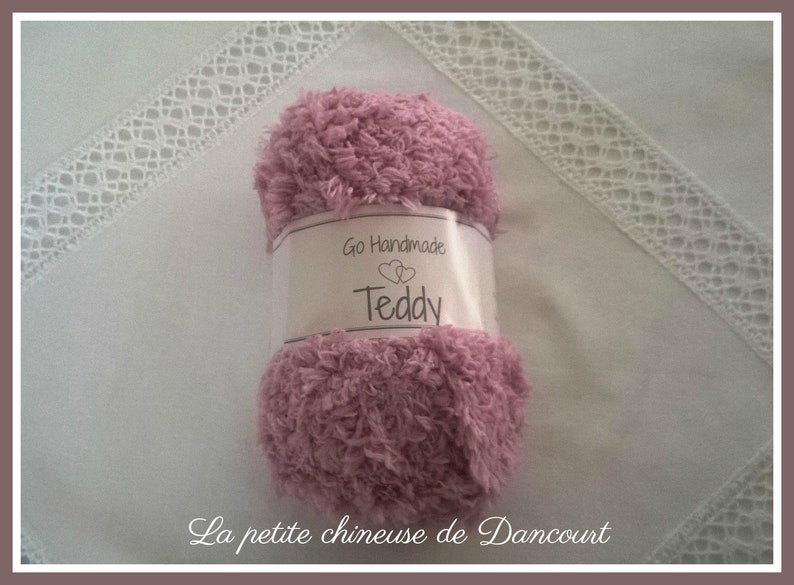 Puede incluir: Un ovillo de lana rosa con la etiqueta "Go Handmade Teddy" sobre una superficie blanca con ribete de encaje blanco.