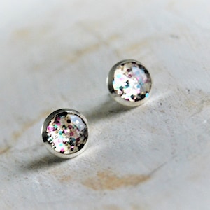 Può includere: Un paio di orecchini a perno argentati con glitter iridescente incorporato nella resina rotonda e trasparente.