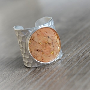 May include: A silver ring with a hammered texture and a circular, cork-like centre. The ring has a wide band and a natural, earthy aesthetic. The cork-like centre has flecks of gold. The ring is a piece of jewellery.