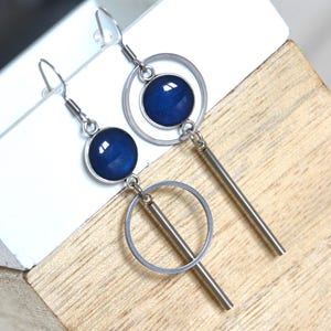 Peut inclure: Une paire de boucles d'oreilles pendantes avec un cadre circulaire argenté et une barre verticale argentée. Chaque boucle d'oreille comprend une pierre cabochon bleu profond ronde sertie dans une monture circulaire argentée, suspendue à un crochet.