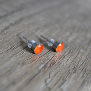 May include: A pair of small stud earrings with a bright orange, circular design. The earrings have a silver-coloured metal base and post. The earrings are displayed on a weathered, grey wooden surface.