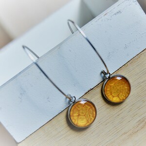 May include: A pair of dangle earrings with a silver-tone metal finish. Each earring features a round, golden-yellow pendant with a textured pattern. The earrings are displayed on a white and light brown wooden surface.