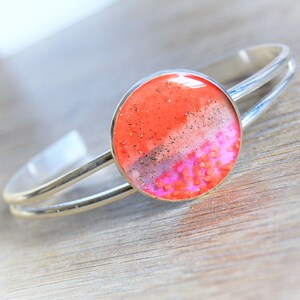May include: A silver bangle bracelet with a round, orange, pink, and white resin cabochon. The cabochon has a glittery finish.