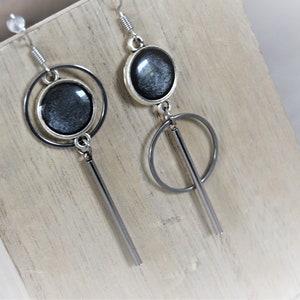 Op de afbeelding: Een paar zilveren oorbellen met een zwarte ronde hanger en een zilveren cirkel- en staafhanger.