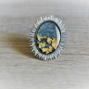 Könnte beinhalten: Ein silberner Ring mit einem blauen und goldenen Harzeinlege. Das Harz hat eine strukturierte, marmorierte Optik mit eingebetteten goldenen Flocken. Der Ring ist in einer silbernen Fassung mit einem Sternendesign gefasst.