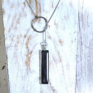Peut inclure: Un collier en chaîne argent avec un pendentif rectangulaire noir et un charme en forme d'anneau argenté.