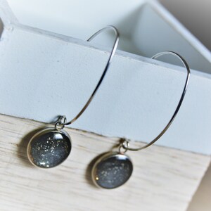 May include: Silver hoop earrings with a round, dark gray pendant filled with sparkling glitter. The earrings are displayed on a white and light wood surface, creating a simple, elegant presentation.