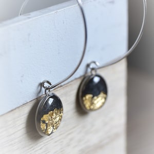 May include: Silver hoop earrings with black and gold oval pendants. The pendants have a dark background with gold leaf accents, creating a striking contrast. The earrings are displayed against a light-colored background.