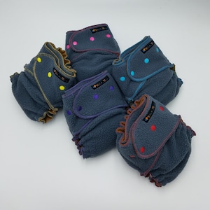 May include: Six grey fleece baby cloth nappies with different coloured trim and poppers. The nappies are folded and stacked on a white surface.