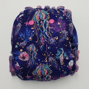 Può includere: Un pannolino di stoffa riutilizzabile blu e viola con una stampa di meduse e cavallucci marini. Il pannolino ha bottoni a pressione rosa e un bordo arricciato.