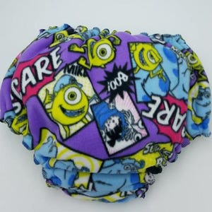 Può includere: Un pannolino per bambini viola e blu con una colorata stampa a cartoni animati con personaggi del film "Monsters, Inc.". Il pannolino ha un bordo a balze ed è decorato con le parole "SCARE" e "BOO!"
