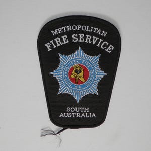 May include: A black embroidered patch featuring the text Metropolitan Fire Service and South Australia in white. The centre displays a blue star emblem with a red circle containing a yellow helmet and the text Metropolitan Fire Service SA.