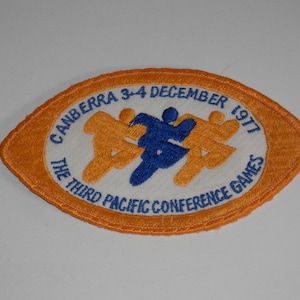 May include: An oval-shaped embroidered patch with an orange border and a white centre. It features two orange figures and one blue figure running, with the text CANBERRA 3-4 DECEMBER 1977 and THE THIRD PACIFIC CONFERENCE GAMES in blue lettering.