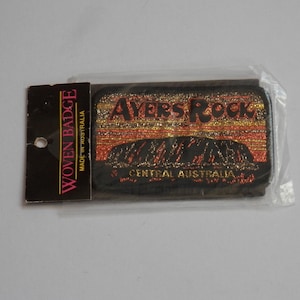 May include: A rectangular woven patch featuring a depiction of Ayers Rock in Central Australia. The design includes metallic gold and orange threads against a black background, with the text AYERS ROCK and CENTRAL AUSTRALIA embroidered.