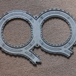 May include: A grey plastic toy train track piece featuring two circular loops connected in the centre. The track has moulded details including cross ties and rail textures, with two protruding connector ends at the bottom for attaching to other tracks.