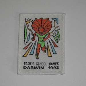 May include: A rectangular white embroidered patch featuring a central orange frilled lizard character running in a green shirt. The design includes colourful geometric accents and the black text "PACIFIC SCHOOL GAMES DARWIN 1992" printed at the bottom.