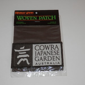 May include: A rectangular woven patch in a clear plastic display sleeve with a black header card labelled Murray Views Woven Patch. The patch features a dark brown background with a white Japanese kanji character and the text Cowra Japanese Garden Australia.