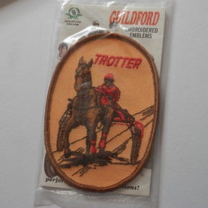 May include: An oval embroidered patch featuring a brown horse pulling a sulky with a driver in red gear. The background is light orange with a dark brown border. The word TROTTER is stitched in red above the horse. It is in a clear plastic package.