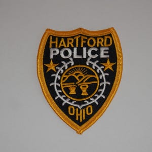 May include: A shield-shaped embroidered patch with a black background and gold border. It features the text HARTFORD POLICE in white, two gold stars, and a central circular emblem depicting a sunrise over hills and wheat in white and gold, with OHIO below.