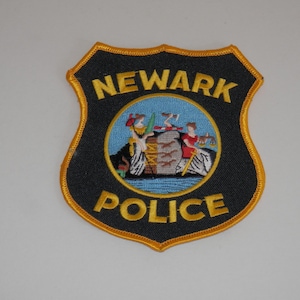 May include: A shield-shaped Newark Police patch with a black background and gold embroidered border. The text NEWARK is at the top and POLICE is at the bottom in gold lettering. A central circular emblem features two figures and a shield design.