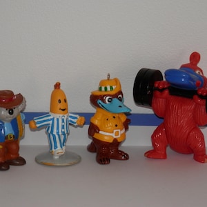 May include: Four small plastic figurines including a koala wearing a brown hat and blue vest, a banana character in blue and white striped pyjamas, a brown platypus in a yellow outfit with a green striped hat, and a red creature lifting a black barbell.